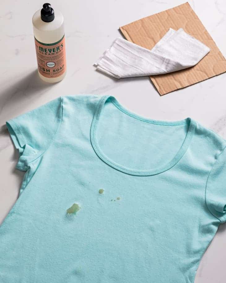 Una mancha de aceite en una camiseta azul, rodeada de materiales de limpieza como jabón para platos, un paño y un trozo de cartón